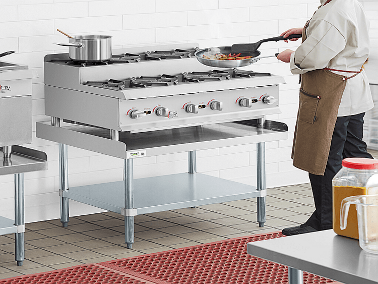 Chef using sautee pan on two tier gas range top sitting on atop a stainless steel prep table with under table shelf