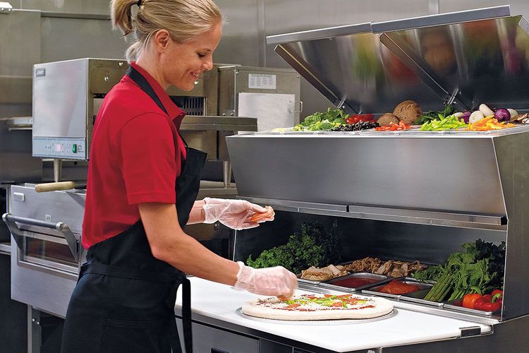 Woman wearing red shirt placing ingredients on a pizza dough round at a Pizza Prep Table