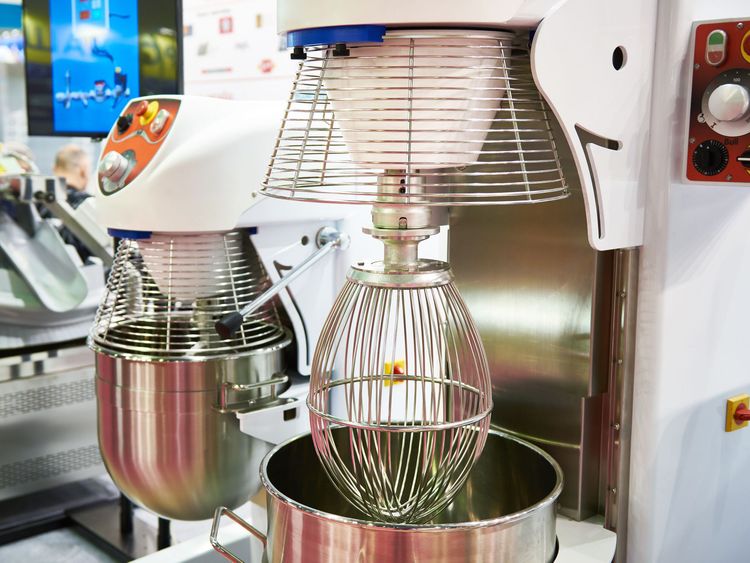 Two planetary mixers side by side. Both with protective cages. One has whisked lowered; the other raised